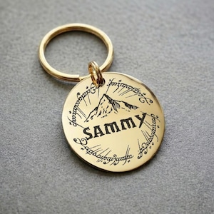 May include: A gold-colored metal keychain with a round pendant. The pendant features a mountain illustration, the name "SAMMY" in bold letters, and script around the edge. A gold ring connects the pendant to the keychain.