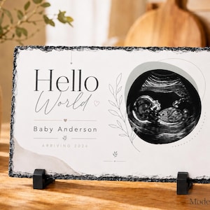 May include: A rectangular slate plaque with a rough, chiseled edge featuring a black and white ultrasound image. The text reads Hello World, Baby Anderson, and Arriving 2026. The design includes thin line art of leaves and small hearts on a white background.