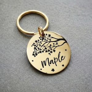 Könnte beinhalten: Ein goldfarbener, runder Hundeanhänger mit einem Astdesign und dem Namen "Maple" in Schreibschrift. Der Anhänger enthält einen goldfarbenen Schlüsselring.