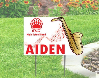 May include: White yard sign with red text and a red paw print. The text reads "El Paso High School Band" and "AIDEN". A gold saxophone with musical notes is in the background.