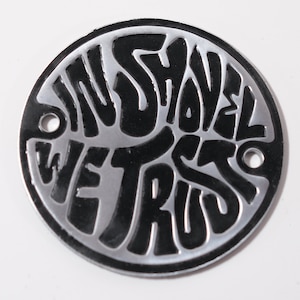 Può includere: Un disco rotondo in metallo argentato con scritta nera che recita "In Shovel We Trust".