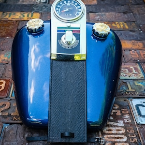 Könnte beinhalten: Ein blauer Motorrad-Benzintank mit einem schwarzen Ledersitz und einem verchromten Tachometer mit der Aufschrift "000000" und "RPM". Der Tank hat zwei verchromte Kappen mit Adlerlogos.