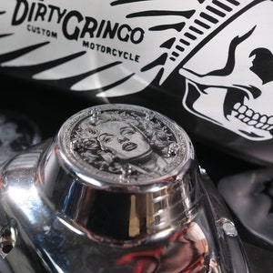 May include: A chrome motorcycle engine cover featuring a detailed engraved image of a woman's face. The cover is decorated with small screws around the edge. The cover is resting on a black motorcycle fuel tank with a white skull and feather design and the text "Dirty Gringo Custom Motorcycle".