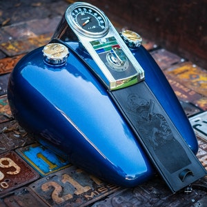 Peut inclure: Un r&eacute;servoir de moto bleu brillant avec un compteur de vitesse chrom&eacute; et un bouchon de r&eacute;servoir. Une sangle en cuir noir avec un motif grav&eacute; est attach&eacute;e. Le r&eacute;servoir repose sur une surface de plaques d'immatriculation vintage.