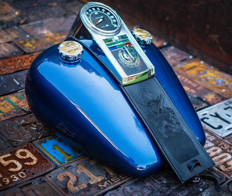 Peut inclure: Un r&eacute;servoir de moto bleu brillant avec un compteur de vitesse chrom&eacute; et un bouchon de r&eacute;servoir. Une sangle en cuir noir avec un motif grav&eacute; est attach&eacute;e. Le r&eacute;servoir repose sur une surface de plaques d'immatriculation vintage.