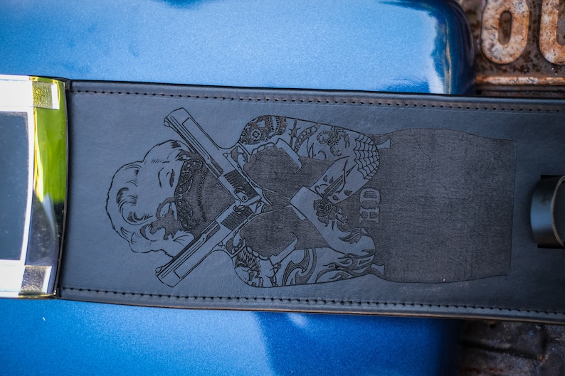 Peut inclure: Ceinture en cuir noir avec un motif grav&eacute; d'une femme avec des pistolets crois&eacute;s et des tatouages. La ceinture a une boucle sur le c&ocirc;t&eacute; gauche. Le design est grav&eacute; dans le cuir.