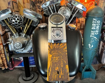 May include: A close-up of a motorcycle engine with chrome accents and a black leather seat with a Native American design. The engine is mounted on a black stand and there is a blue and white bomb-shaped object in the background with the text "The Dirty Gringo" on it.