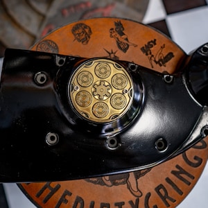 Op de afbeelding: Een zwarte motorfiets motorbehuizing met een ronde, gouden decoratieve plaat met de woorden "Special" en "38 Special".