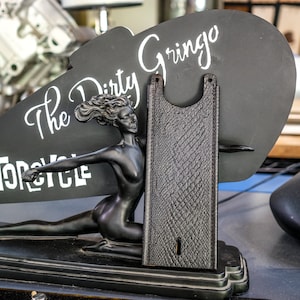 Puede incluir: Un asiento de moto negro con un diseño de cuero texturizado. El asiento se muestra en un soporte negro con una figura esculpida de una mujer y un letrero que dice "The Dirty Gringo Motorcycle".