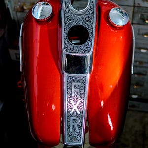 Puede incluir: Primer plano de un depósito de gasolina de motocicleta en un tono rojo vibrante. El depósito presenta un panel decorativo plateado con motivos florales y las letras "FXR". Los detalles cromados incluyen dos tapones de combustible circulares.