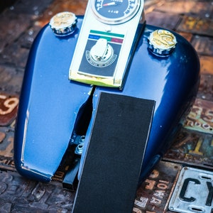 Peut inclure: Un r&eacute;servoir d'essence de moto bleu avec un compteur de vitesse, une jauge de carburant et deux bouchons de r&eacute;servoir chrom&eacute;s. Le r&eacute;servoir a un coussin rectangulaire noir et une sangle noire. Le r&eacute;servoir est sur une surface faite de plaques d'immatriculation.