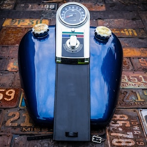 Op de afbeelding: Een blauwe motorfiets benzinetank met een chromen snelheidsmeter en bedieningselementen. De tank heeft twee chromen doppen aan de bovenkant en een zwart leren zadel.