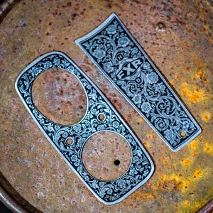 Puede incluir: Dos paneles decorativos de metal con intrincados diseños florales y de volutas. Un panel tiene dos aberturas circulares, el otro tiene las letras "FXR" dentro de un rombo. Los paneles son plateados con detalles en negro.