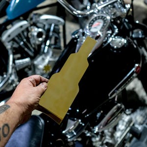May include: A person holding a yellow, bottle-shaped template near a black motorcycle. The motorcycle has chrome handlebars and a speedometer. The person's arm has a tattoo that says "NATO".