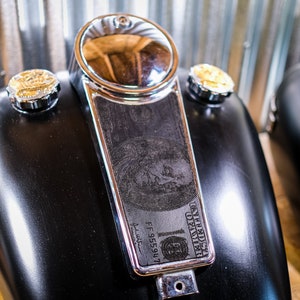Könnte beinhalten: Schwarzer Motorrad-Benzintank mit Chromverkleidung und einem Dollar-Schein-Design an der Seite. Der Benzintank hat zwei verchromte Kappen mit goldenen Akzenten.