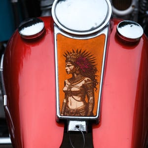 May include: A red motorcycle gas tank with a detailed illustration of a woman in profile, adorned with a feathered headdress and jewelry. The tank has chrome accents and a glossy finish, showcasing a blend of art and machinery.