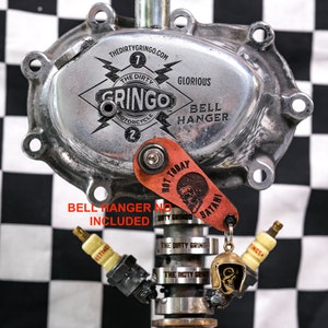 Puede incluir: Un colgador de campana de motocicleta plateado con las palabras "The Dirty Gringo" y "Glorious Bell Hanger" grabadas en &eacute;l. Un llavero de cuero rojo con una calavera y las palabras "Hot Today" y "Satani" est&aacute; unido al colgador de campana.