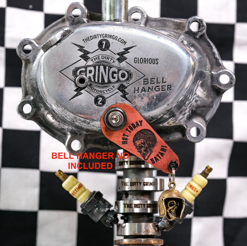 Puede incluir: Un colgador de campana de motocicleta plateado con las palabras "The Dirty Gringo" y "Glorious Bell Hanger" grabadas en &eacute;l. Un llavero de cuero rojo con una calavera y las palabras "Hot Today" y "Satani" est&aacute; unido al colgador de campana.