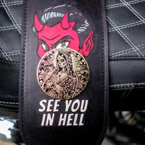 Puede incluir: Correa de cuero negro con una ilustración de diablo rojo y una moneda dorada con un diseño del Día de los Muertos. El texto "SEE YOU IN HELL" está impreso en blanco.