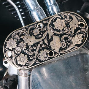 May include: An oval, decorative motorcycle part with a black background and a gold floral design. The design includes flowers and leaves. The part has several holes and is mounted on a motorcycle.
