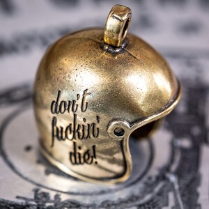 Puede incluir: Un amuleto de metal de color dorado en forma de casco de f&uacute;tbol americano con las palabras "don't fuckin' die!" grabadas en el lateral.