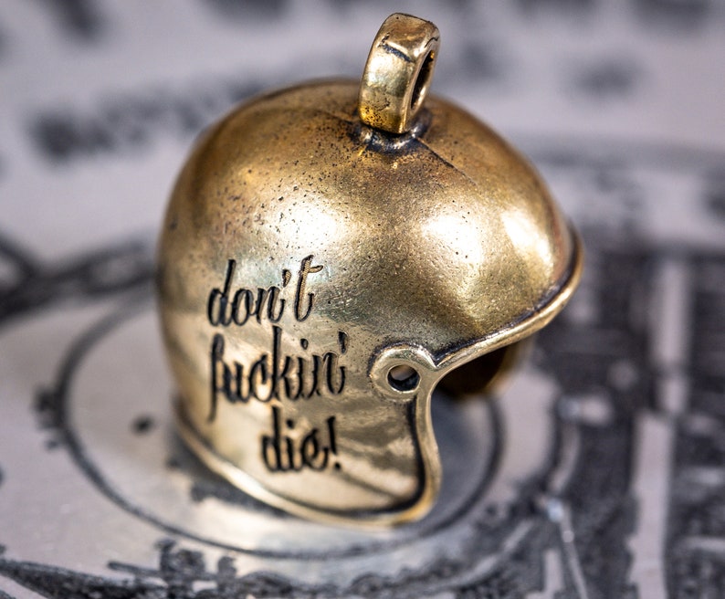 Puede incluir: Un amuleto de metal de color dorado en forma de casco de f&uacute;tbol americano con las palabras "don't fuckin' die!" grabadas en el lateral.