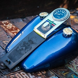 Peut inclure: Un r&eacute;servoir d'essence de moto bleu avec un compteur de vitesse et des d&eacute;tails chrom&eacute;s. Une sangle en cuir noir avec un motif grav&eacute; est attach&eacute;e. Le r&eacute;servoir a deux bouchons de r&eacute;servoir chrom&eacute;s. Le fond est compos&eacute; de plaques d'immatriculation vintage.