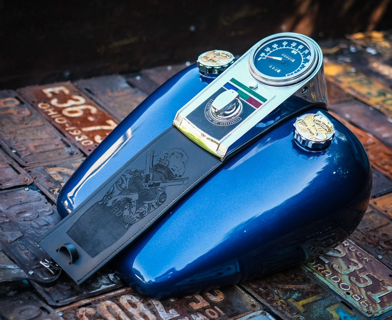 Peut inclure: Un r&eacute;servoir d'essence de moto bleu avec un compteur de vitesse et des d&eacute;tails chrom&eacute;s. Une sangle en cuir noir avec un motif grav&eacute; est attach&eacute;e. Le r&eacute;servoir a deux bouchons de r&eacute;servoir chrom&eacute;s. Le fond est compos&eacute; de plaques d'immatriculation vintage.