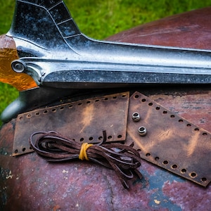 Puede incluir: Dos correas de cuero marrón con agujeros perforados alrededor de los bordes, un cordón de cuero marrón y dos remaches de metal plateado sobre una superficie de metal oxidado.