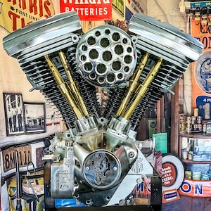 Puede incluir: Primer plano de un motor de motocicleta cromado pulido con empujadores de color dorado. El motor tiene un filtro de aire grande y redondo con una cubierta cromada y múltiples orificios. El motor está montado en un soporte negro y está rodeado de varios letreros y otros objetos.