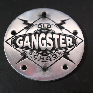 Könnte beinhalten: Ein rundes Metallemblem in Silber mit einer schwarzen Raute in der Mitte. Die Raute enthält die Worte "Old School Gangster". Die Raute ist von zwei Blitzen umgeben.