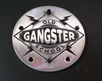 May include: A silver round metal emblem with a black diamond shape in the center. The diamond has the words "Old School Gangster" inside. The diamond is surrounded by two lightning bolts.