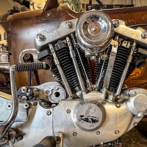 Pode incluir: Close-up de um motor de motocicleta cromado polido com um design de caveira e ossos cruzados no lado. O motor está montado em uma estrutura marrom enferrujada.