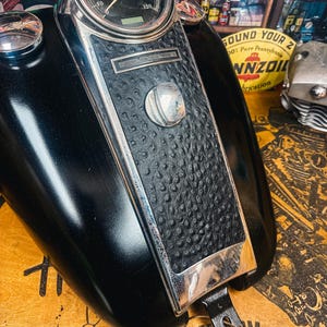May include: Black motorcycle gas tank with a chrome speedometer and a textured leather panel. The speedometer reads up to 120 mph. The tank has a chrome fuel cap and a mounting bracket.