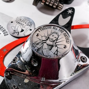 May include: Close-up of motorcycle parts, including a chrome cover with the text "I FORGIVE ALL THE HATERS!". Other elements include a graphic with the text "LOOK OUT!" and a "THE DIRTY GRINGO" custom motorcycle graphic.
