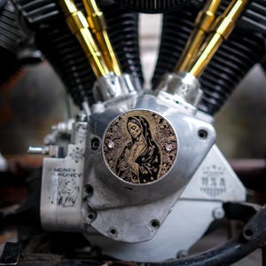 Puede incluir: Primer plano de un motor de motocicleta con tubos dorados y una cubierta decorativa redonda con una figura religiosa. El motor es plateado con el texto "MONEY HONEY" y "USA" visible.