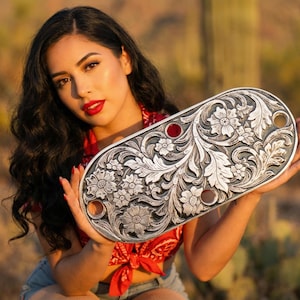 May include: A silver-colored, oval-shaped metal object with intricate floral and leaf carvings. The object has three circular holes and two red accents. The design is detailed and symmetrical, with a vintage aesthetic.