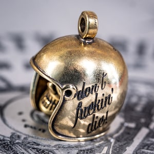 Puede incluir: Un amuleto de casco de metal dorado con las palabras "Don't fuckin' die!" grabadas en el lateral.