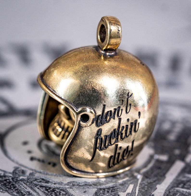 Puede incluir: Un amuleto de casco de metal dorado con las palabras "Don't fuckin' die!" grabadas en el lateral.