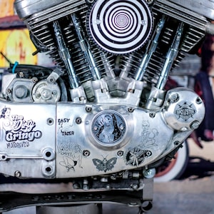Puede incluir: Primer plano de un motor de motocicleta con acabado plateado. El motor presenta un diseño en espiral en blanco y negro y varias calcomanías, incluyendo "The Dirty Gringo Motorcycle". Se ven cactus en el fondo.