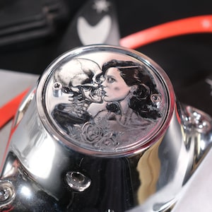 Puede incluir: Una pieza de motocicleta cromada con un diseño grabado de un cráneo y una mujer besándose en la parte superior.
