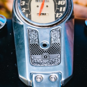 Könnte beinhalten: Nahaufnahme eines Motorrad-Tachometers mit Chrom-Finish. Das Messgerät zeigt Zahlen und einen orangefarbenen Zeiger. Das Chromgehäuse weist verzierte Gravuren und die Worte "Old School Patriot" auf.