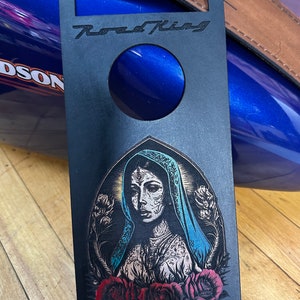 May include: Black motorcycle gas tank cover with a cutout for the gas cap. The cover features a graphic of a woman with a floral crown and a blue veil, surrounded by roses. The text "Road King" is printed at the top of the cover.