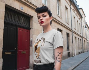 May include: A light gray t-shirt with a graphic of a woman and the words "Dirty Gringo Motorcycle". The woman is wearing a brown swimsuit and high heels. The model is wearing black jeans and has tattoos on her arm.