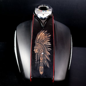 Puede incluir: Depósito de gasolina de motocicleta negro con una franja roja y un grabado detallado de un indio americano con un tocado. El depósito tiene una tapa cromada con la palabra "Indian" en ella.