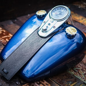 Op de afbeelding: Een blauwe motorfiets benzinetank met een zwarte leren band, een chromen snelheidsmeter en twee gouden benzine doppen.