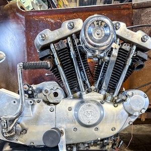 May include: Close-up of a vintage motorcycle engine. The engine is silver with chrome accents, featuring multiple cylinders and a prominent air filter. The background shows a rusty metal surface.