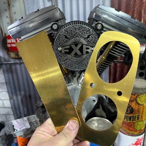 Puede incluir: Dos piezas de motocicleta de metal dorado se sostienen frente a un motor de motocicleta. Una pieza es rectangular y la otra tiene dos recortes circulares. El motor tiene un acabado plateado y un emblema que dice "OLD FXR SCHOOL".