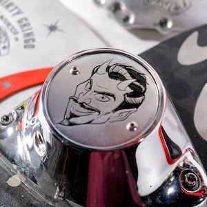 Puede incluir: Una pieza de motocicleta cromada con un grabado detallado de un rostro de diablo. El grabado es en blanco y negro y presenta una cabeza cornuda con una sonrisa traviesa.