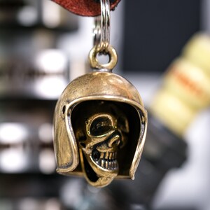 Puede incluir: Un llavero de calavera de color dorado con un dise&ntilde;o de casco. La calavera est&aacute; detallada con cuencas de los ojos, una nariz y una boca. El llavero est&aacute; unido a una correa de cuero.